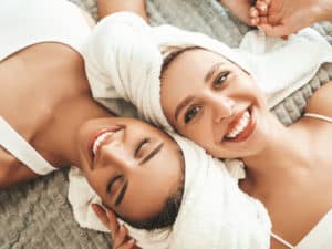 Two young beautiful smiling women in white bathrobes and towels on head.